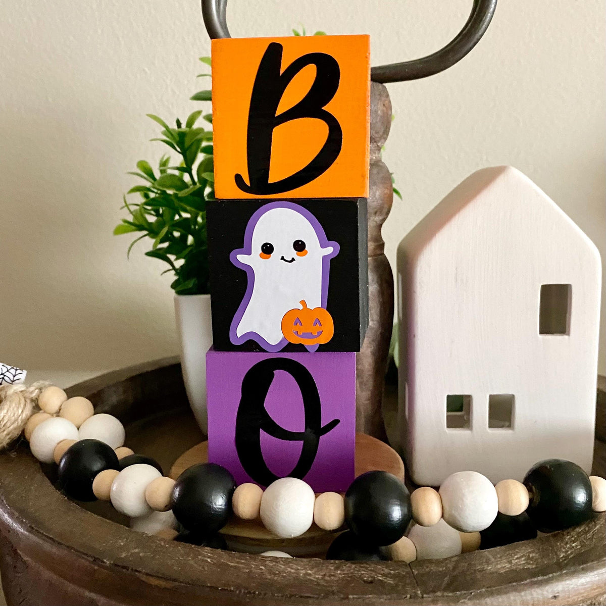 A decorative Halloween block sign spelling BOO, with an orange block showing a black letter B, a black block with a smiling ghost holding a pumpkin, and a purple block with a black letter O.