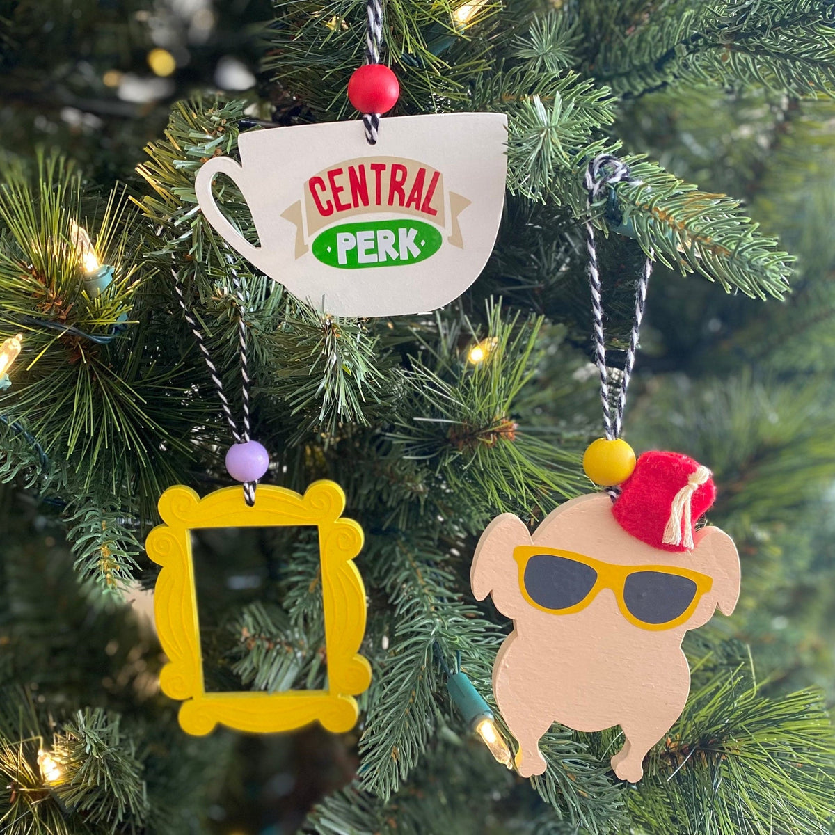 Three colorful Christmas ornaments shaped like a Central Perk teacup, a yellow picture frame, and a character wearing yellow sunglasses and a red Santa hat, hanging on a green Christmas tree with warm string lights.