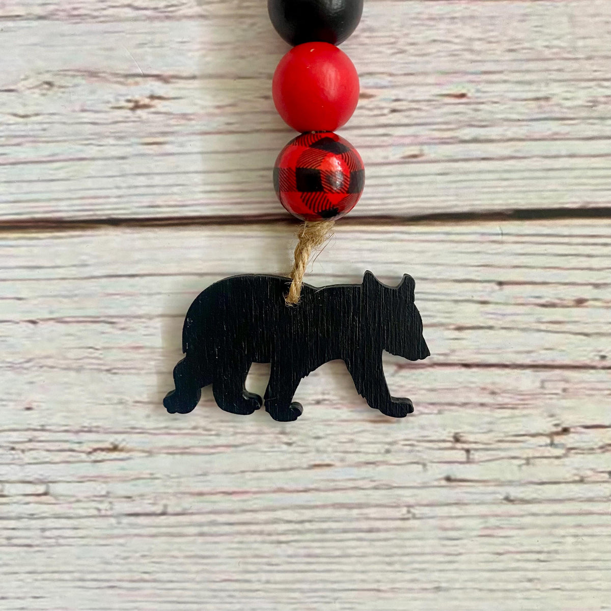 A beaded necklace or ornament featuring a black wooden bear silhouette charm hanging from a natural twine loop, with red and black beads above it, set against a whitewashed wood plank background.