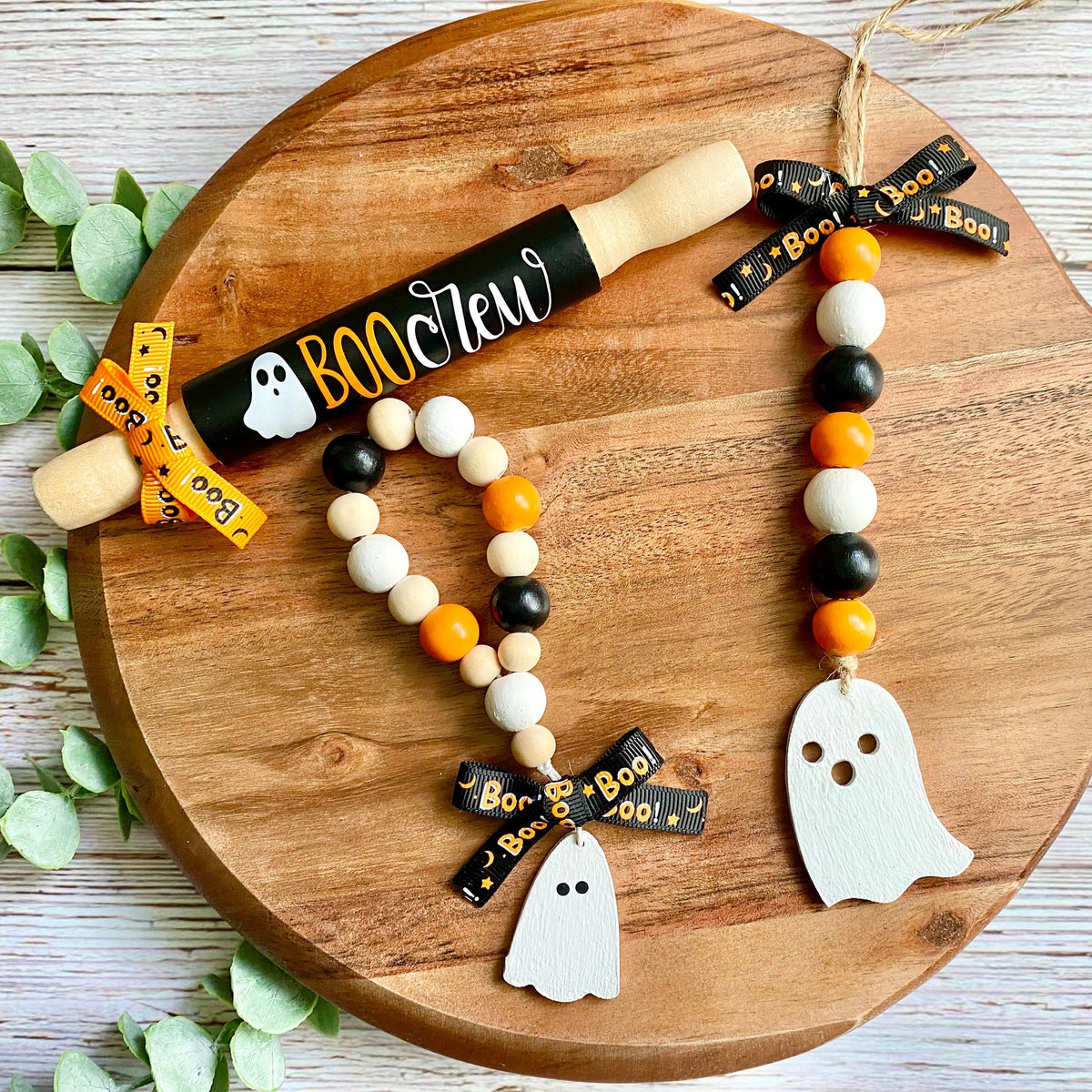 Halloween-themed wooden bead garlands and a matching wooden rolling pin set on a round wooden serving board, with eucalyptus leaves on a white wood background.