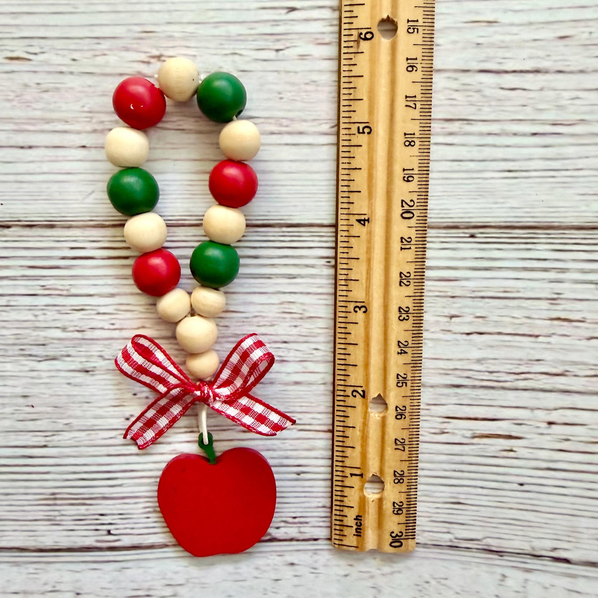A beaded garland with red, green, and cream wooden beads, a red gingham bow, and a red apple ornament, shown next to a wooden ruler on a whitewashed wood background.