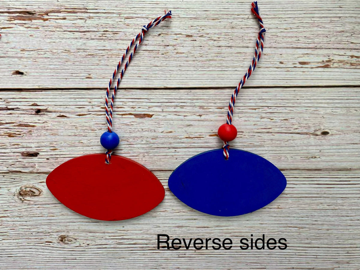 Two football-shaped wooden ornaments in red and blue, each with a braided red, white, and blue hanging cord and a single colored bead, displayed on a rustic white wood background. The text 'Reverse sides' is printed at the bottom.