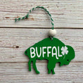 A green wooden St. Patrick's Day ornament shaped like a buffalo, featuring the word 'BUFFALO' in white, a white shamrock, and a hanging loop with a white bead on a rustic white wood background.