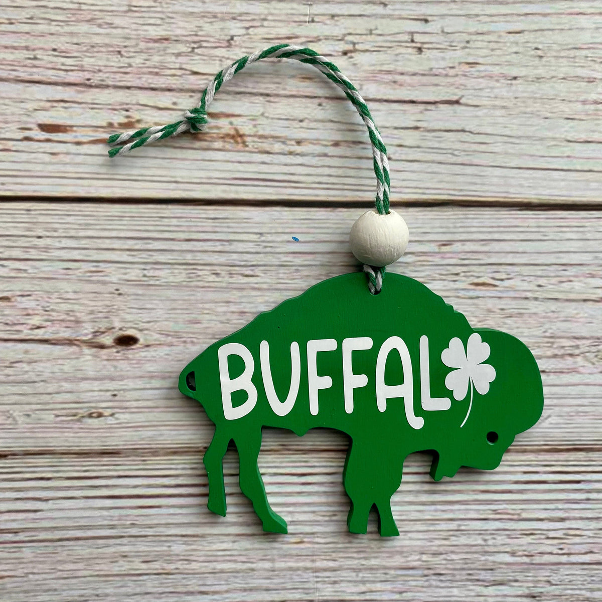 A green wooden St. Patrick's Day ornament shaped like a buffalo, featuring the word 'BUFFALO' in white, a white shamrock, and a hanging loop with a white bead on a rustic white wood background.