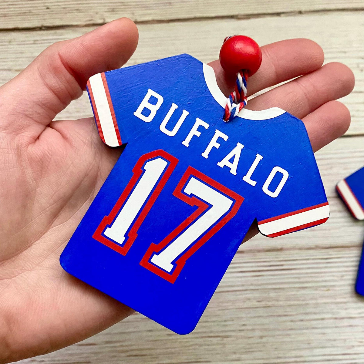 A hand holding a blue Buffalo 17 wooden ornament shaped like a sports jersey, with white lettering and numbers outlined in red, and a small red hanging bead.