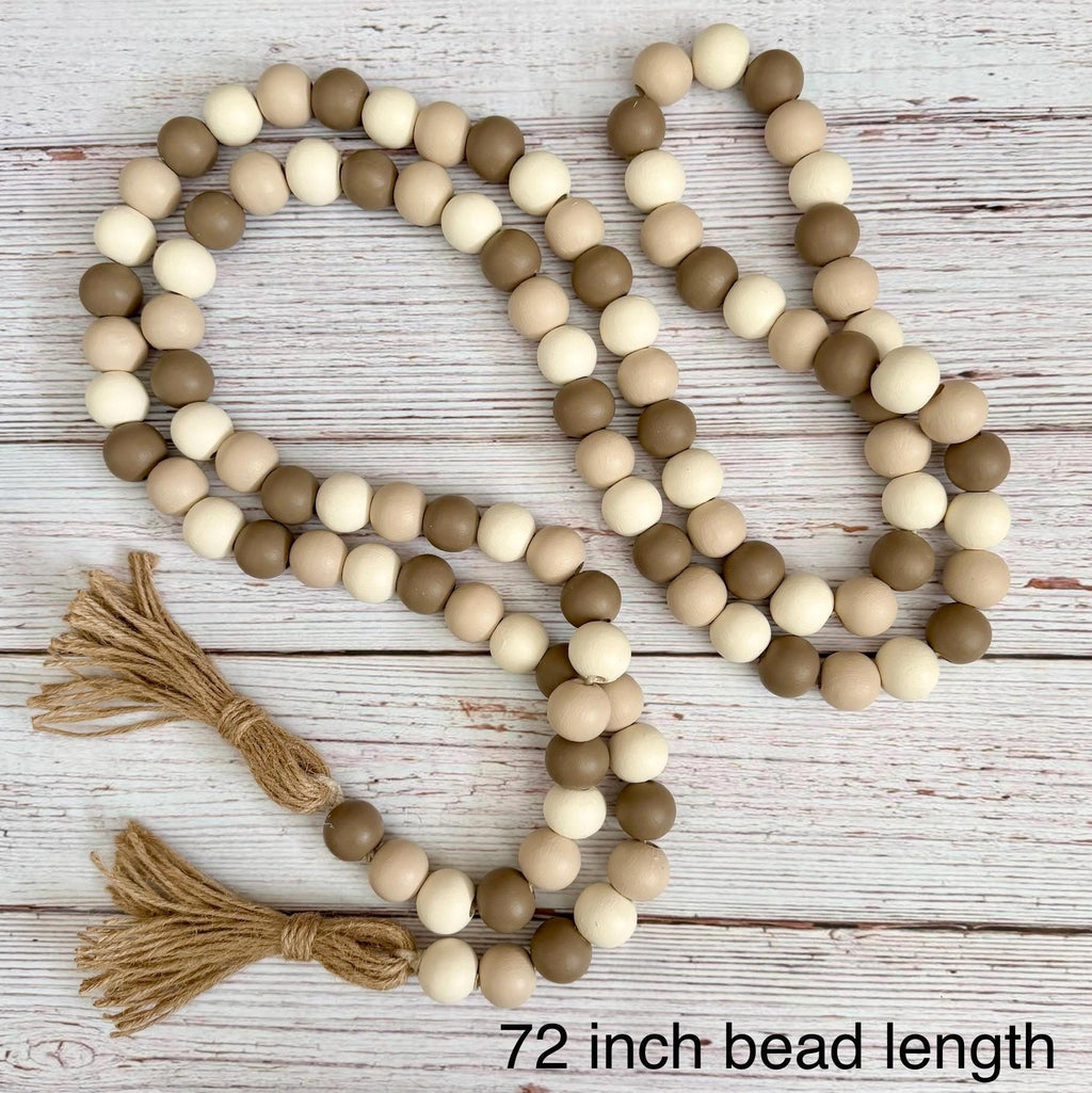 Wooden bead garland with natural brown, beige, and cream beads, featuring two jute tassels, displayed on a white rustic wood plank background with text reading '72 inch bead length'.