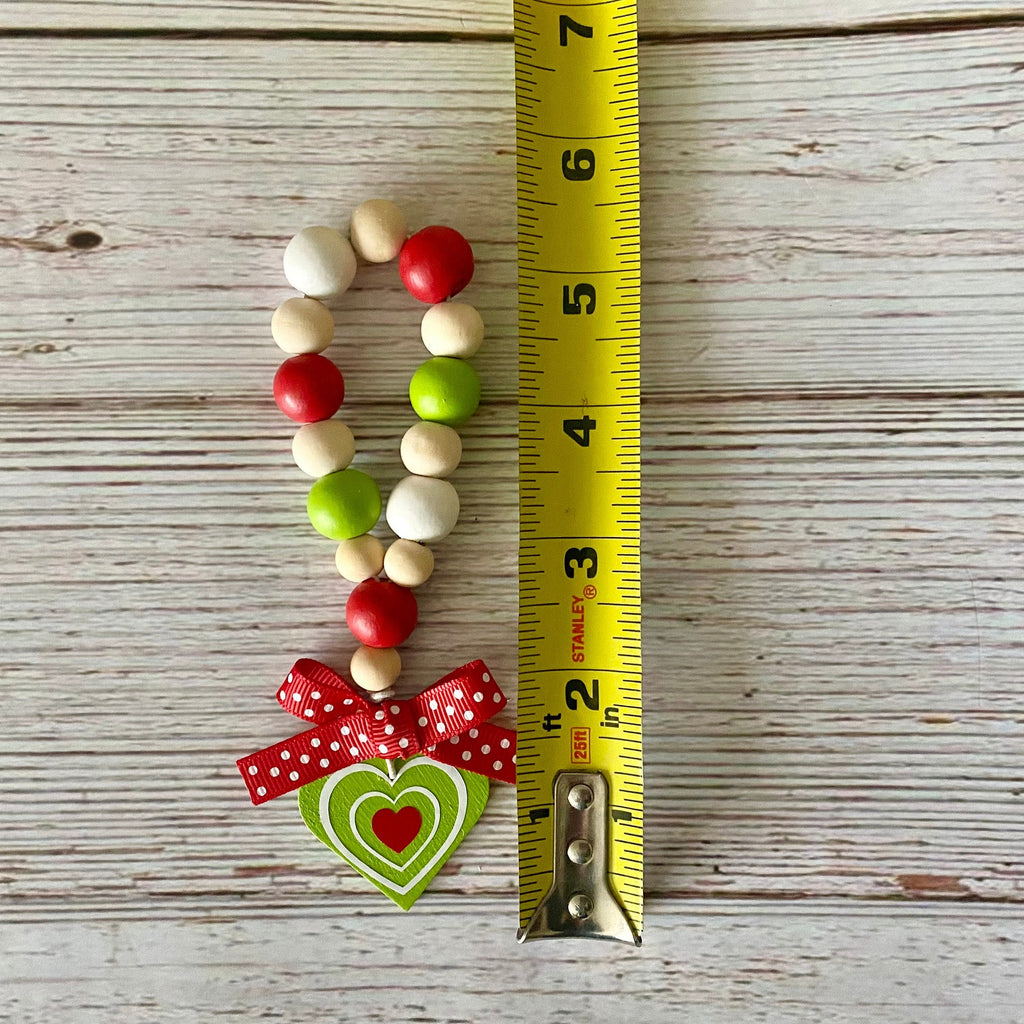A handmade beaded charm or keychain with red, green, and natural wood beads, a red polka dot ribbon bow, and a green heart-shaped metal charm, shown next to a yellow tape measure on a whitewashed wooden surface.