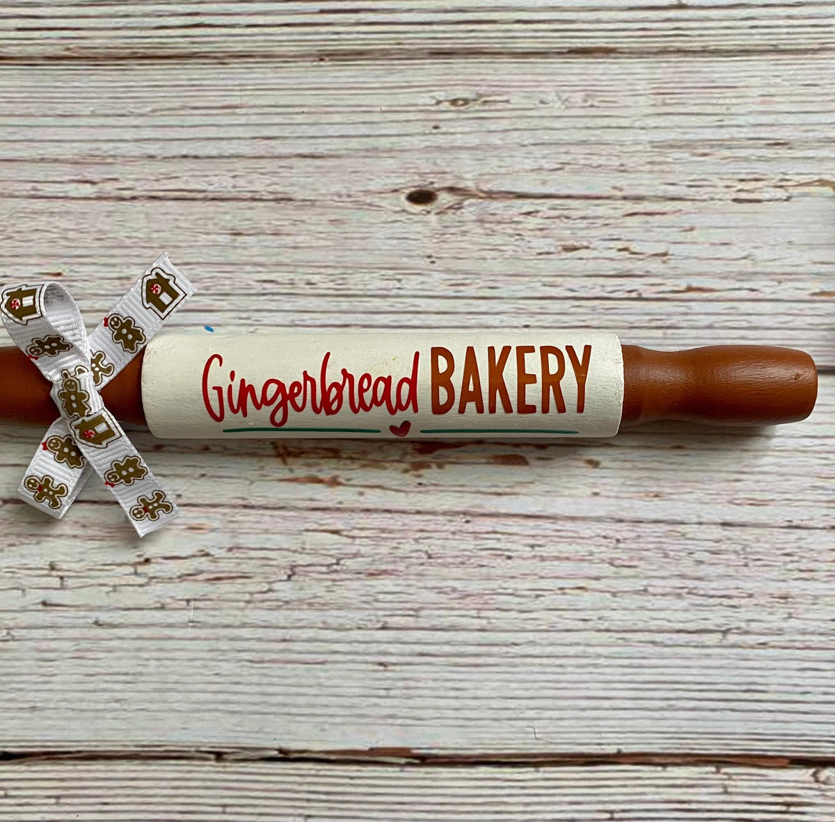 A gingerbread-themed rolling pin with brown wooden ends and a white center wrapped in a white ribbon printed with gingerbread cookies, placed on a white wood plank background next to a decorative bead garland.