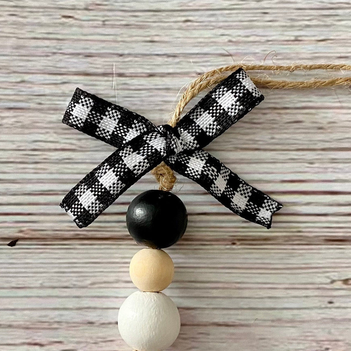 A rustic Christmas ornament featuring a black and white gingham ribbon bow, a black bead, and two natural wood beads on a whitewashed wood background.