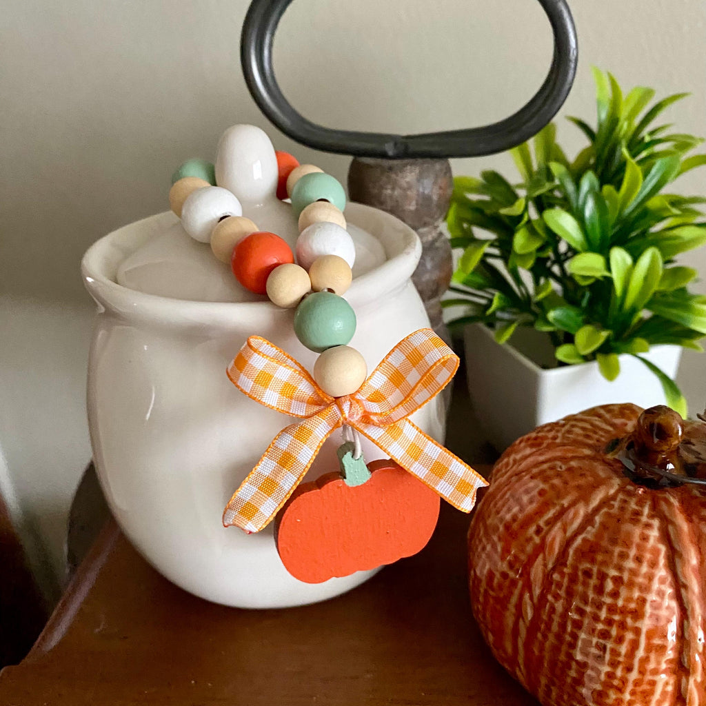 A decorative autumn beaded garland draped over a white ceramic canister, featuring pastel wood beads, an orange gingham ribbon bow, and an orange pumpkin charm, styled on a tabletop with a potted plant and a ceramic pumpkin in the background.