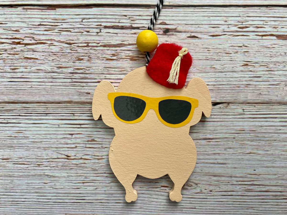 A wooden turkey-shaped ornament or keychain charm with yellow sunglasses, a red felt hat, and a black-and-white hanging cord, photographed on a rustic whitewashed wood surface.