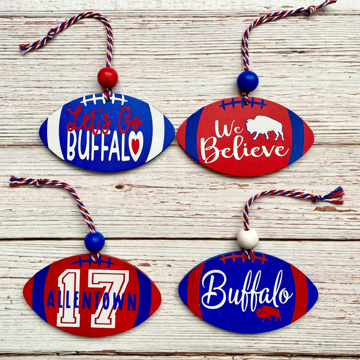 Four football-shaped wooden ornaments with red, white, and blue painted stripes and sports-themed designs, displayed on a light wood plank background. The ornaments feature phrases like 'Let's Go BUFFALO', 'We Believe', 'ALENTOWN', and 'Buffalo', with hanging cords and decorative beads.