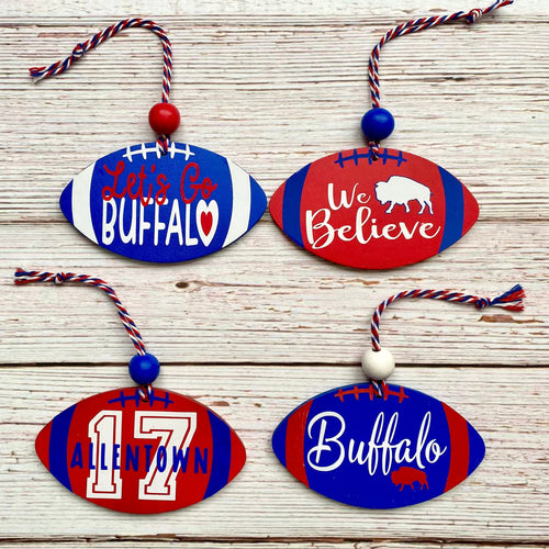 Four football-shaped wooden ornaments with red, white, and blue painted stripes and sports-themed designs, displayed on a light wood plank background. The ornaments feature phrases like 'Let's Go BUFFALO', 'We Believe', 'ALENTOWN', and 'Buffalo', with hanging cords and decorative beads.