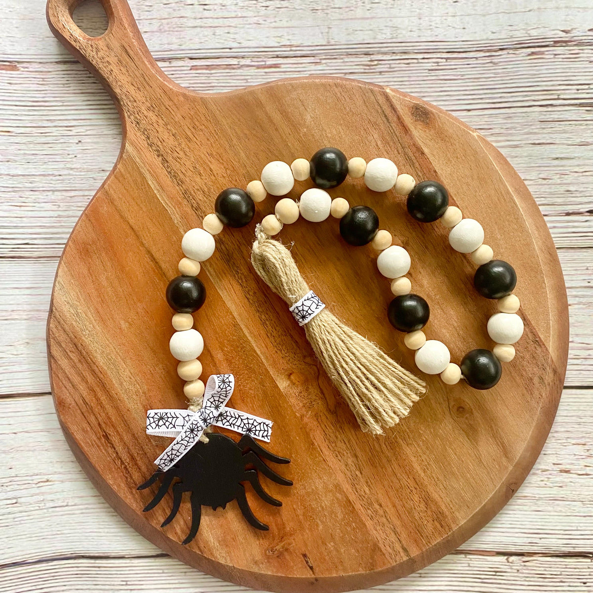 A beaded garland with alternating black and cream beads, a beige jute tassel, a silver rhinestone bow, and a black spider silhouette charm, displayed on a round wooden cutting board.