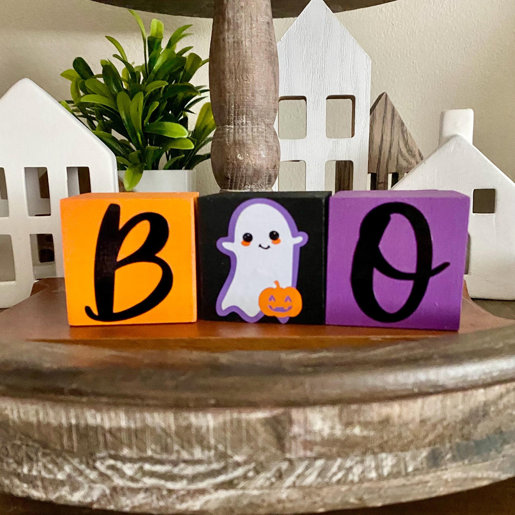Three wooden block letters spelling 'BOO' for Halloween, with a black 'B' on an orange block, a smiling white ghost holding a pumpkin on a black block, and a purple 'O' with a black outline, displayed on a rustic wooden surface.