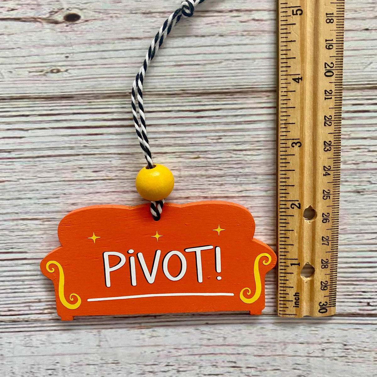 An orange couch  shaped ornament with the word 'PIVOT!' in white, decorated with yellow scroll accents and a black-and-white twisted string, shown next to a wooden ruler on a whitewashed wood background.
