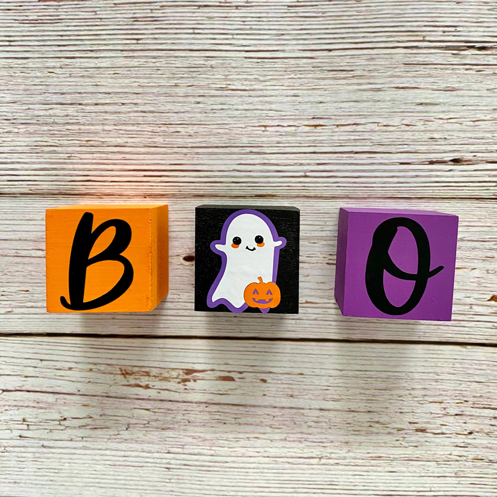 Three decorative wooden blocks spelling 'BOO' on a whitewashed wood background. The left block is orange with a black letter 'B', the middle block is black with a white ghost and orange pumpkin, and the right block is purple with a black letter 'O'.