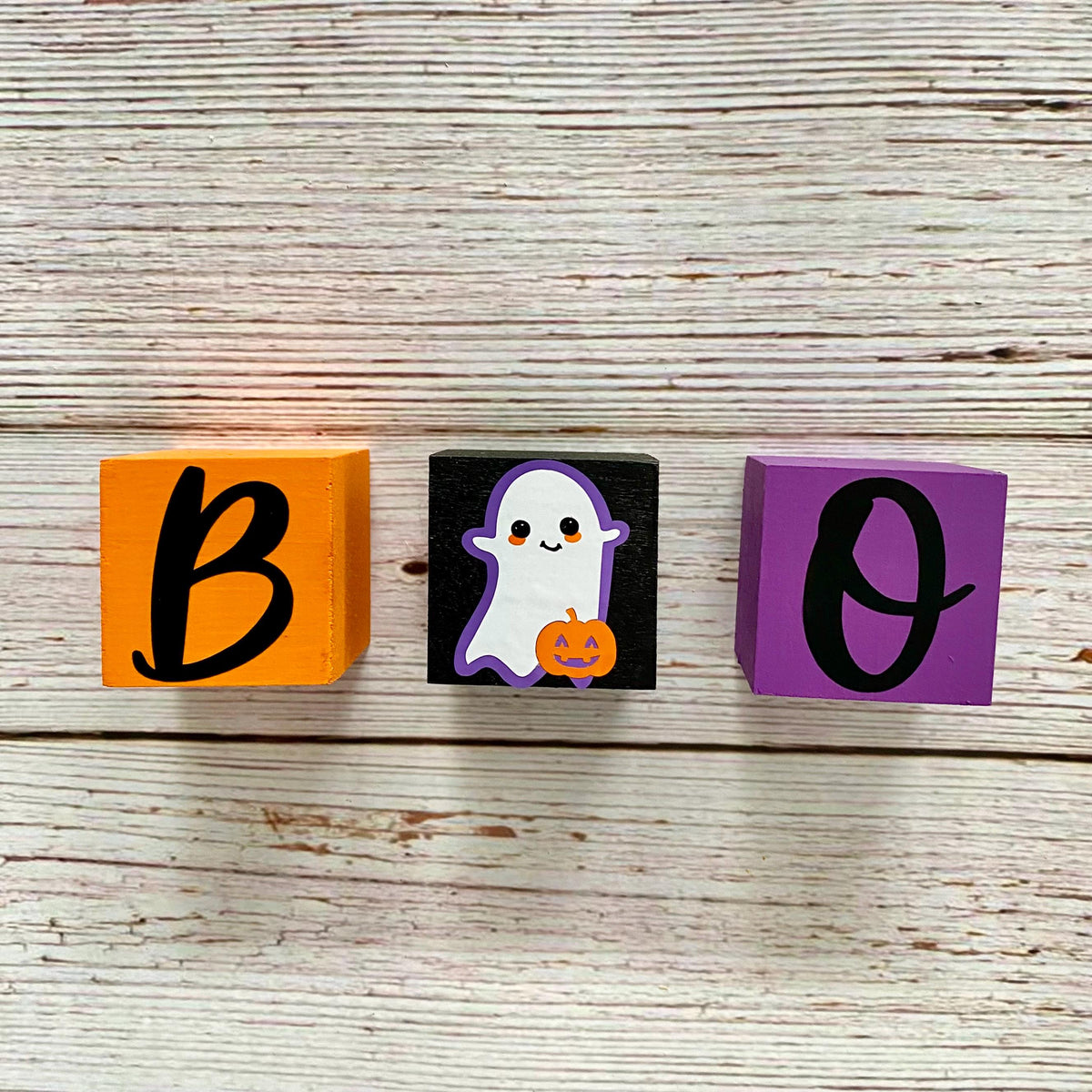 Three decorative wooden blocks spelling 'BOO' on a whitewashed wood background. The left block is orange with a black letter 'B', the middle block is black with a white ghost and orange pumpkin, and the right block is purple with a black letter 'O'.