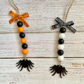 Two Halloween bead garlands with black spider cutouts, displayed on a whitewashed wooden surface. One garland features orange and black beads with an orange spiderweb ribbon bow, and the other features black and white beads with a black and white spiderweb ribbon bow.