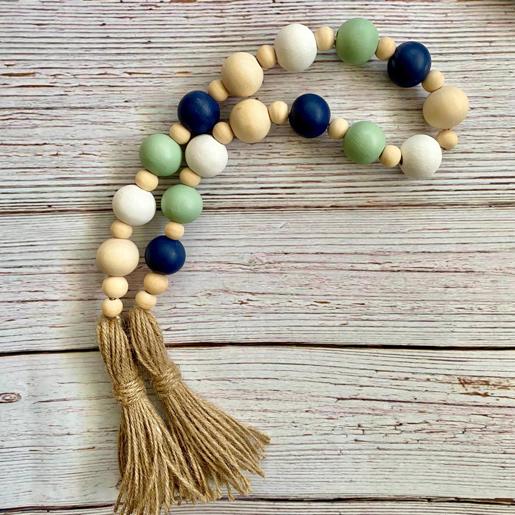 A beaded baby necklace or garland with natural wood beads, painted green and navy blue beads, white beads, and two tan jute tassels, displayed on a rustic whitewashed wooden surface.