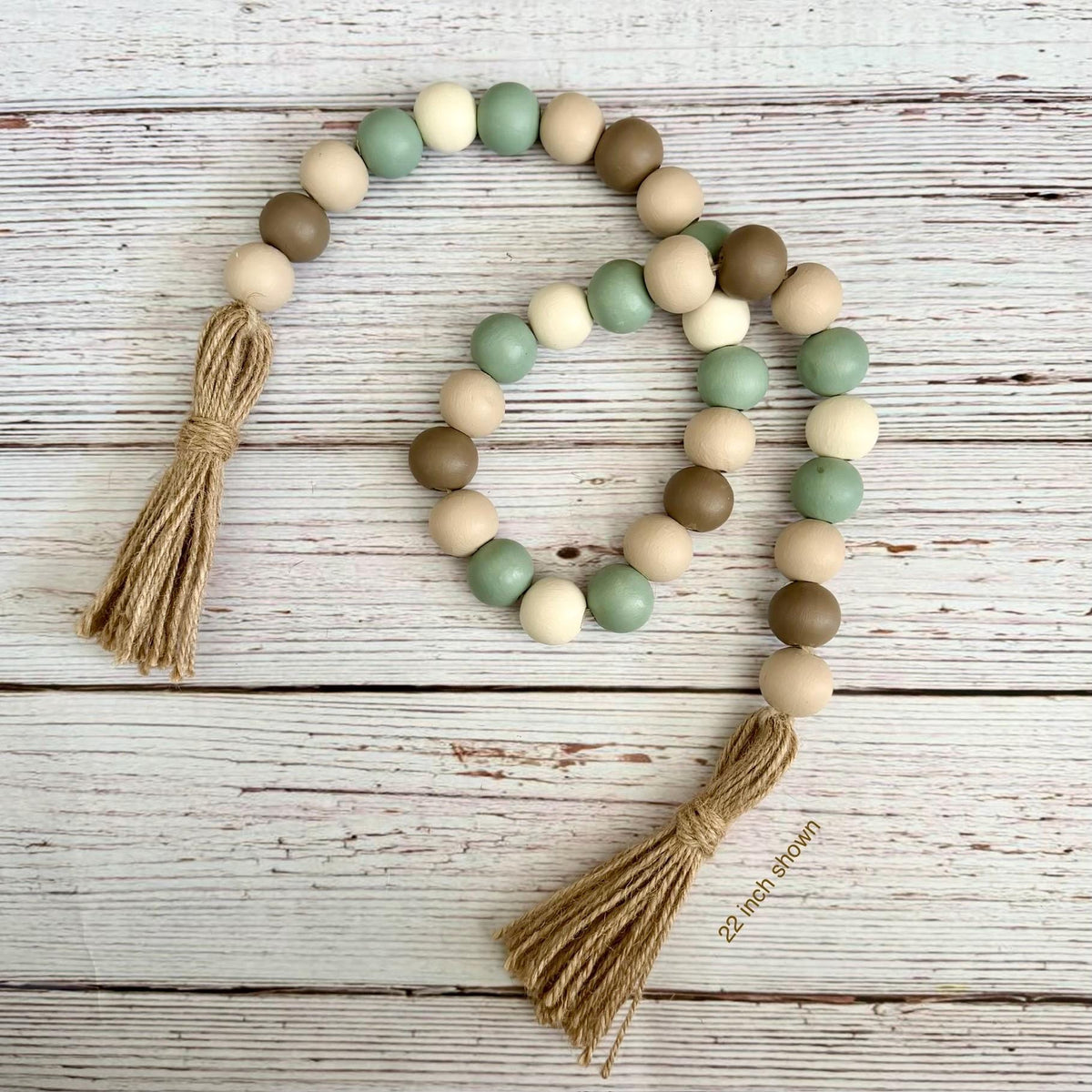 A wooden bead garland with two jute tassels, featuring matte beads in sage green, light beige, and medium brown, arranged on a white-washed wooden background.
