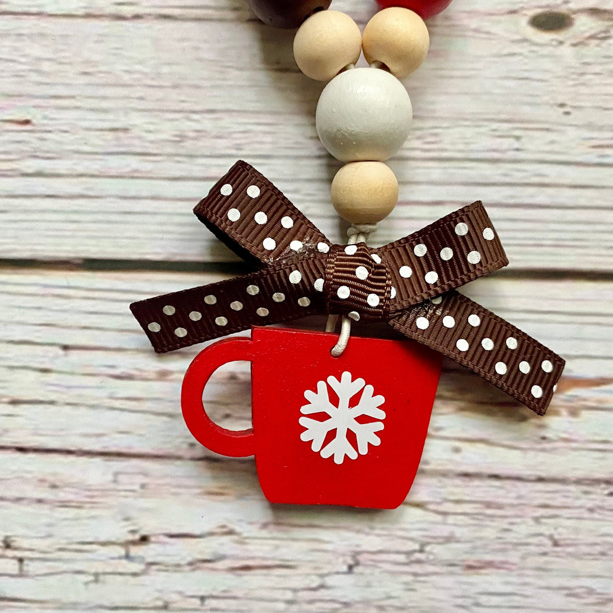 A close-up of a holiday-themed beaded necklace or garland featuring a red mug charm with a white snowflake, tied with a brown polka-dot ribbon, on a whitewashed wood background.