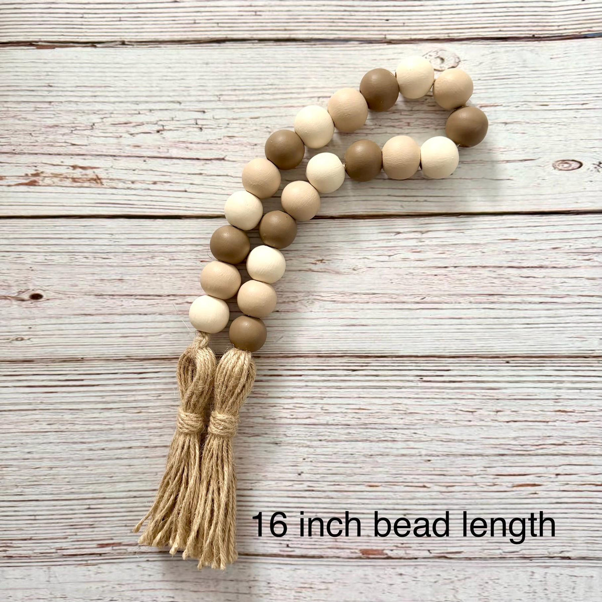 A beaded garland with alternating light beige, medium brown, and cream wooden beads, finished with a natural jute tassel, displayed on a whitewashed wood background with text reading '16 inch bead length'.