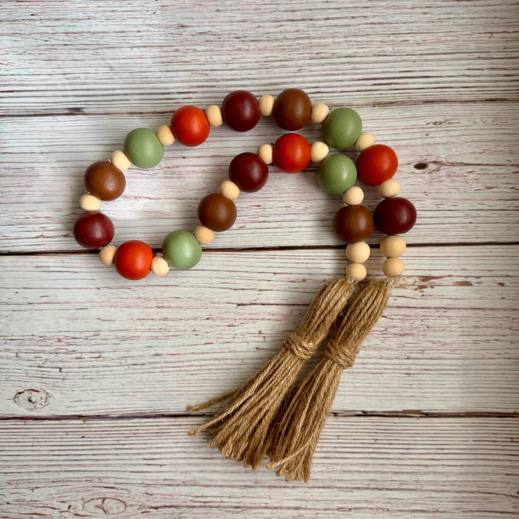A wooden bead garland with two braided jute tassels, featuring alternating beads in dark red, orange, sage green, medium brown, and natural light wood, displayed on a rustic white-washed wooden background.