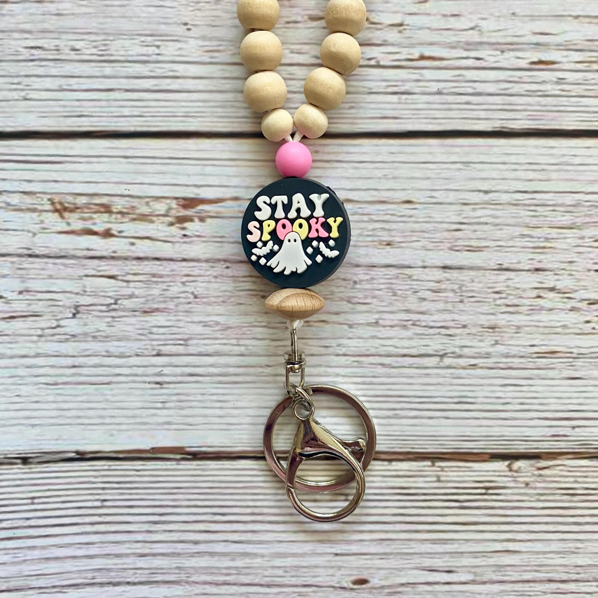 A beaded keychain or bag charm with natural wood beads, a pink accent bead, a round black 'STAY SPOOKY' charm with a white ghost, and a metal key ring, displayed on a whitewashed wood background.
