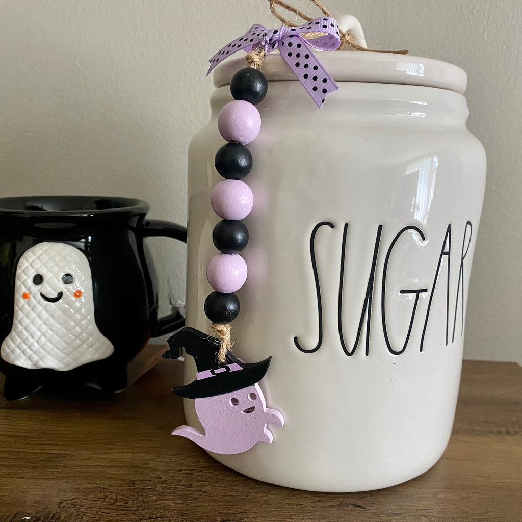 A white ceramic sugar canister with 'SUGAR' written on the front, decorated with a purple and black bead garland and a ghost charm, displayed next to a black mug with a white ghost on a wooden table.