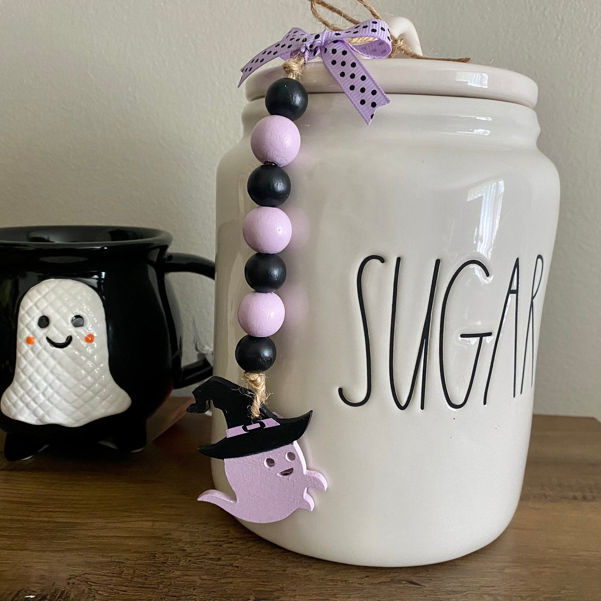 A white ceramic sugar canister with 'SUGAR' written on the front, decorated with a purple and black bead garland and a ghost charm, displayed next to a black mug with a white ghost on a wooden table.
