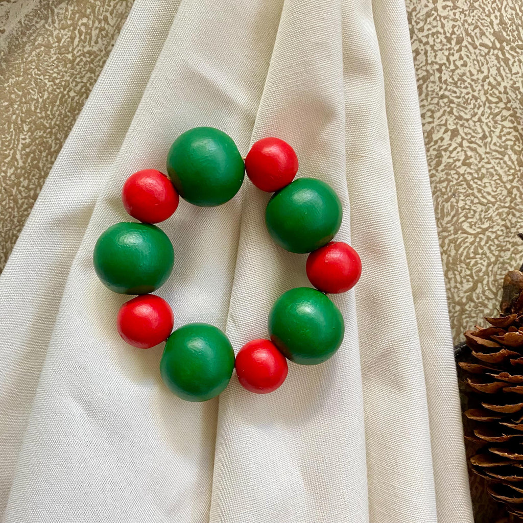 A beaded napkin ring made of alternating green and red wooden beads, displayed on a draped cream fabric background.