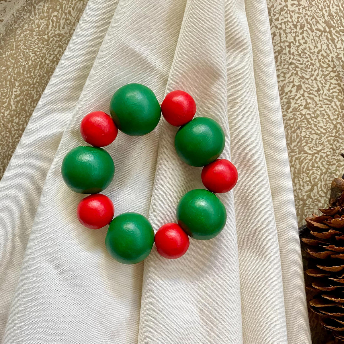 A beaded napkin ring made of alternating green and red wooden beads, displayed on a draped cream fabric background.