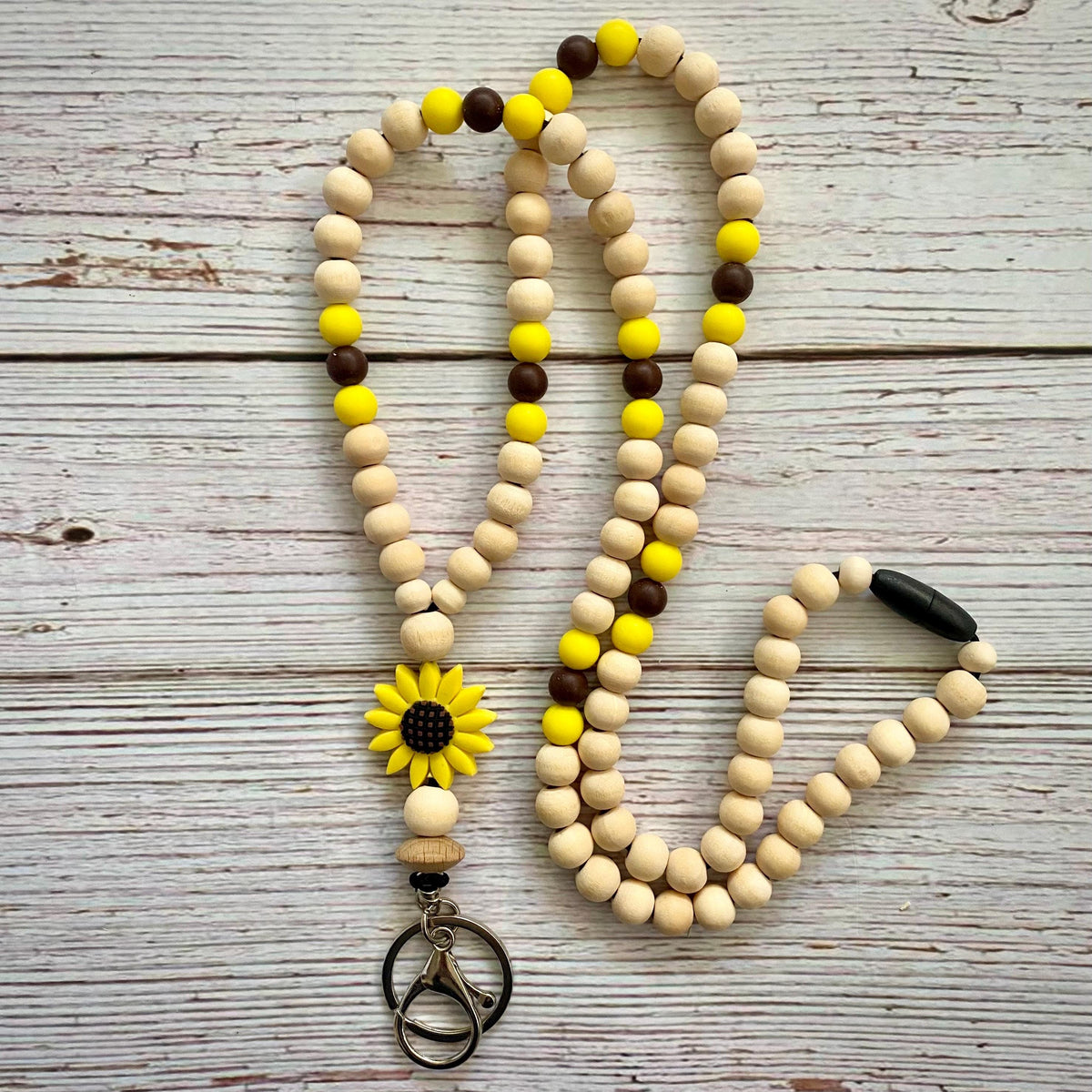 A wooden bead necklace laid flat on a whitewashed wood surface. The necklace is made of natural light wood beads with alternating bright yellow and dark brown beads, a yellow sunflower charm with a black center, and a black oval bead near one end.