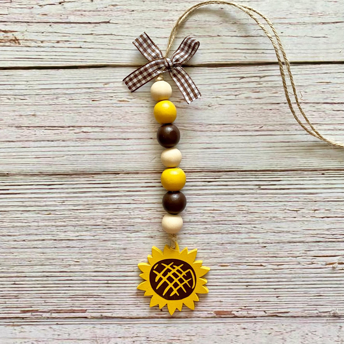 A rustic wooden bead hanging ornament with a brown and beige gingham ribbon bow, yellow and brown wooden beads, and a yellow sunflower-shaped charm, displayed on a whitewashed wooden background.
