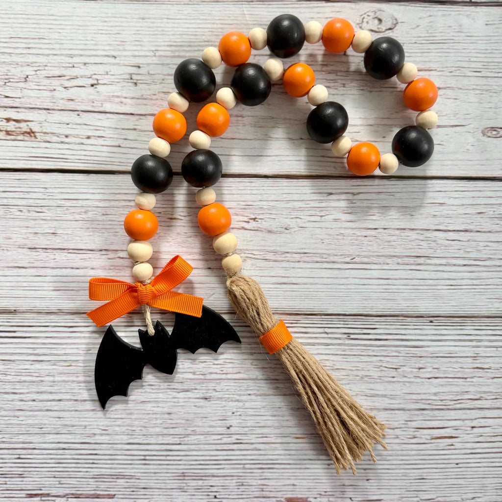 A wooden bead garland with alternating black, orange, and natural beige beads, finished with a jute tassel wrapped in orange ribbon and a black bat decoration tied with an orange bow, displayed on a whitewashed wood background.
