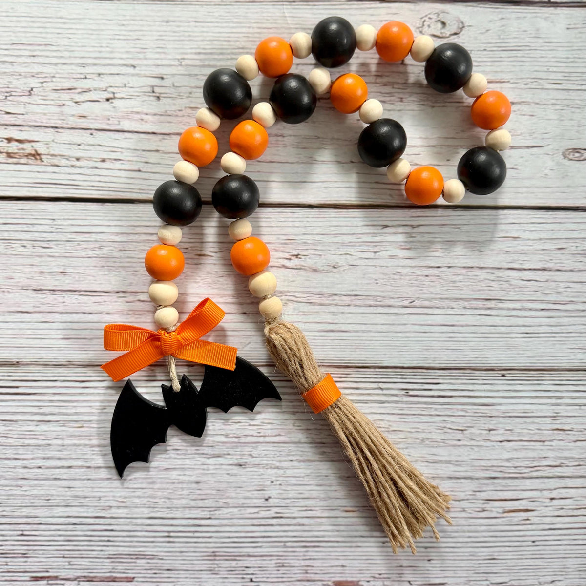 A wooden bead garland with alternating black, orange, and natural beige beads, finished with a jute tassel wrapped in orange ribbon and a black bat decoration tied with an orange bow, displayed on a whitewashed wood background.