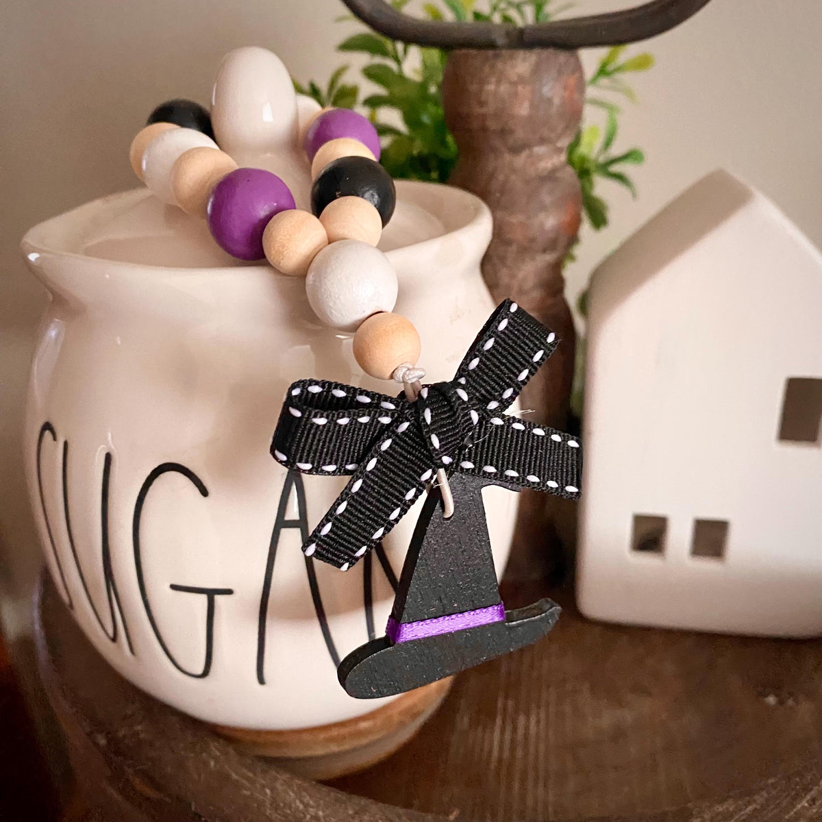 A decorative beaded garland with purple, black, white, and natural wood beads, tied around a white ceramic canister and featuring a black witch hat charm with a purple band and black-and-white ribbon bow, styled on a wooden surface with a white house decor piece in the background.