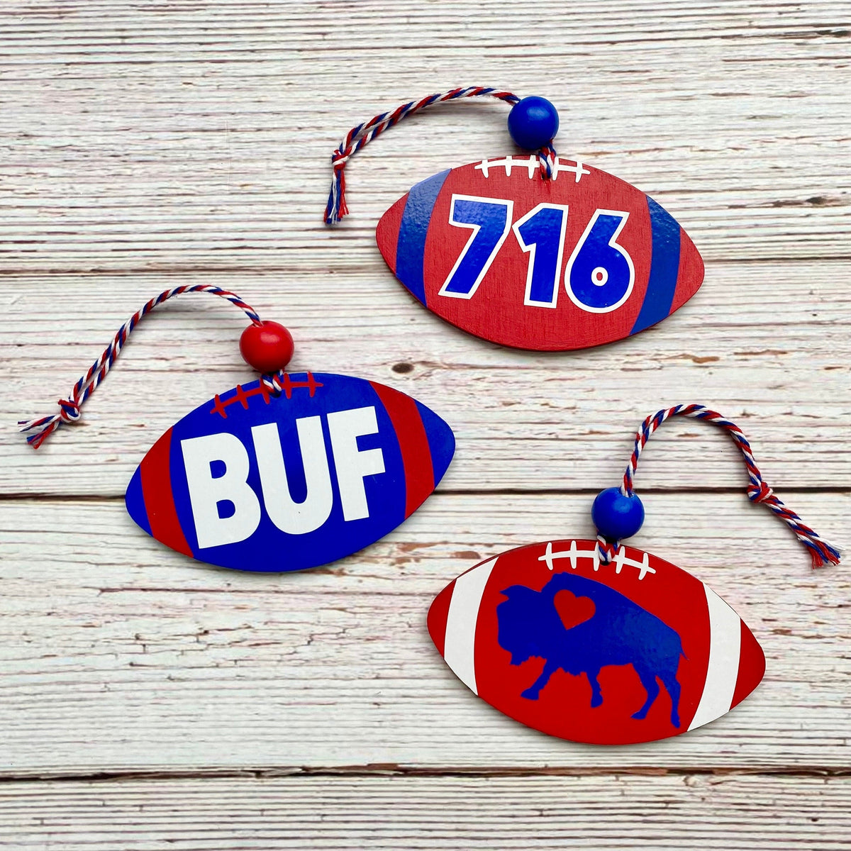 Three wooden football-shaped ornaments with red and blue paint, displayed on a light wood background. One ornament features the letters 'BUF' in white, another has the number '716' in blue, and the third shows a blue buffalo silhouette.
