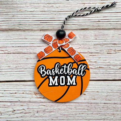 A circular orange basketball-shaped ornament with the words 'Basketball MOM' in white and black script, decorated with a small orange-and-white basketball-patterned bow and a black-and-white string, displayed on a rustic white wood background.