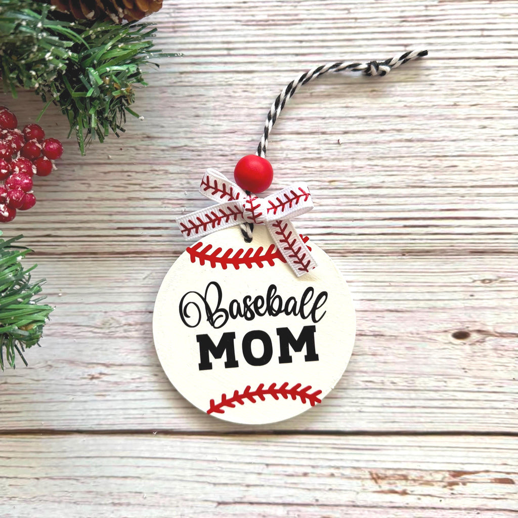 A round wooden baseball ornament with 'Baseball MOM' printed in black script and block lettering, decorated with red baseball seam details, a red bead, and a ribbon loop, displayed on a rustic whitewashed wood surface with holiday greenery and red berries as props.