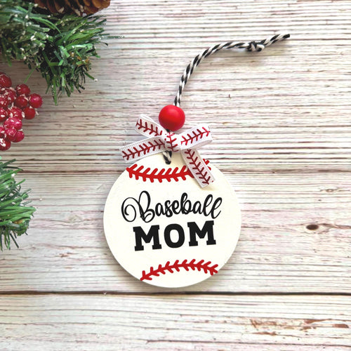 A round wooden baseball ornament with 'Baseball MOM' printed in black script and block lettering, decorated with red baseball seam details, a red bead, and a ribbon loop, displayed on a rustic whitewashed wood surface with holiday greenery and red berries as props.