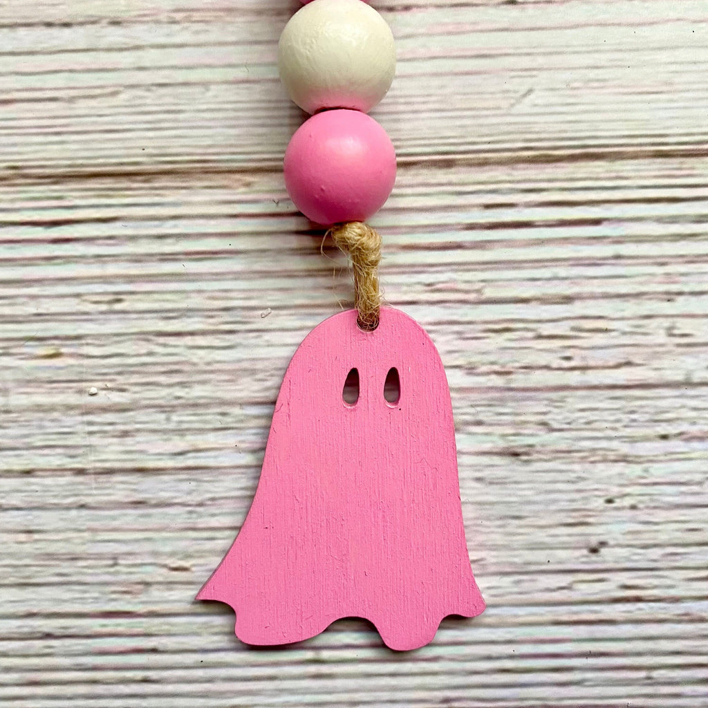 A pink wooden ghost ornament hanging from a string with pink and natural wood beads, set against a rustic whitewashed wooden background.
