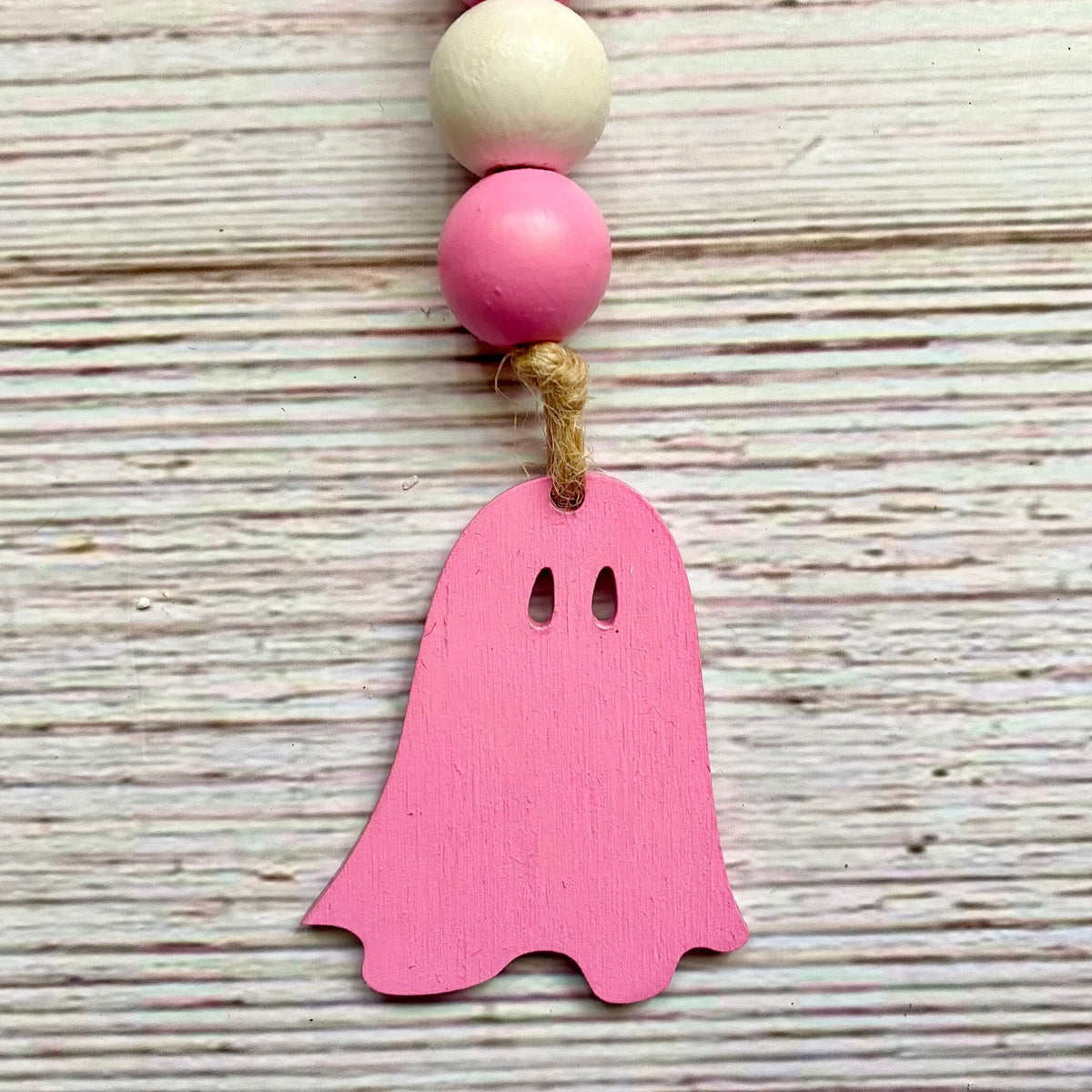 A pink wooden ghost ornament hanging from a string with pink and natural wood beads, set against a rustic whitewashed wooden background.