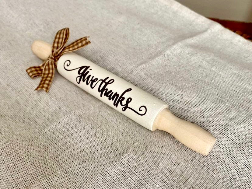 A white wooden rolling pin with 'give thanks' written in black script, tied with a brown and beige plaid ribbon, resting on a textured beige linen fabric.