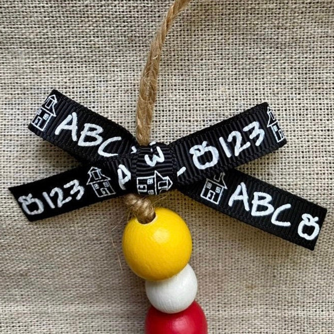 A school-themed hanging ornament featuring a black ribbon bow printed with white letters, numbers, and house icons, suspended from a natural jute string. The ornament is decorated with three wooden beads in yellow, white, and red, set against a textured beige burlap background.
