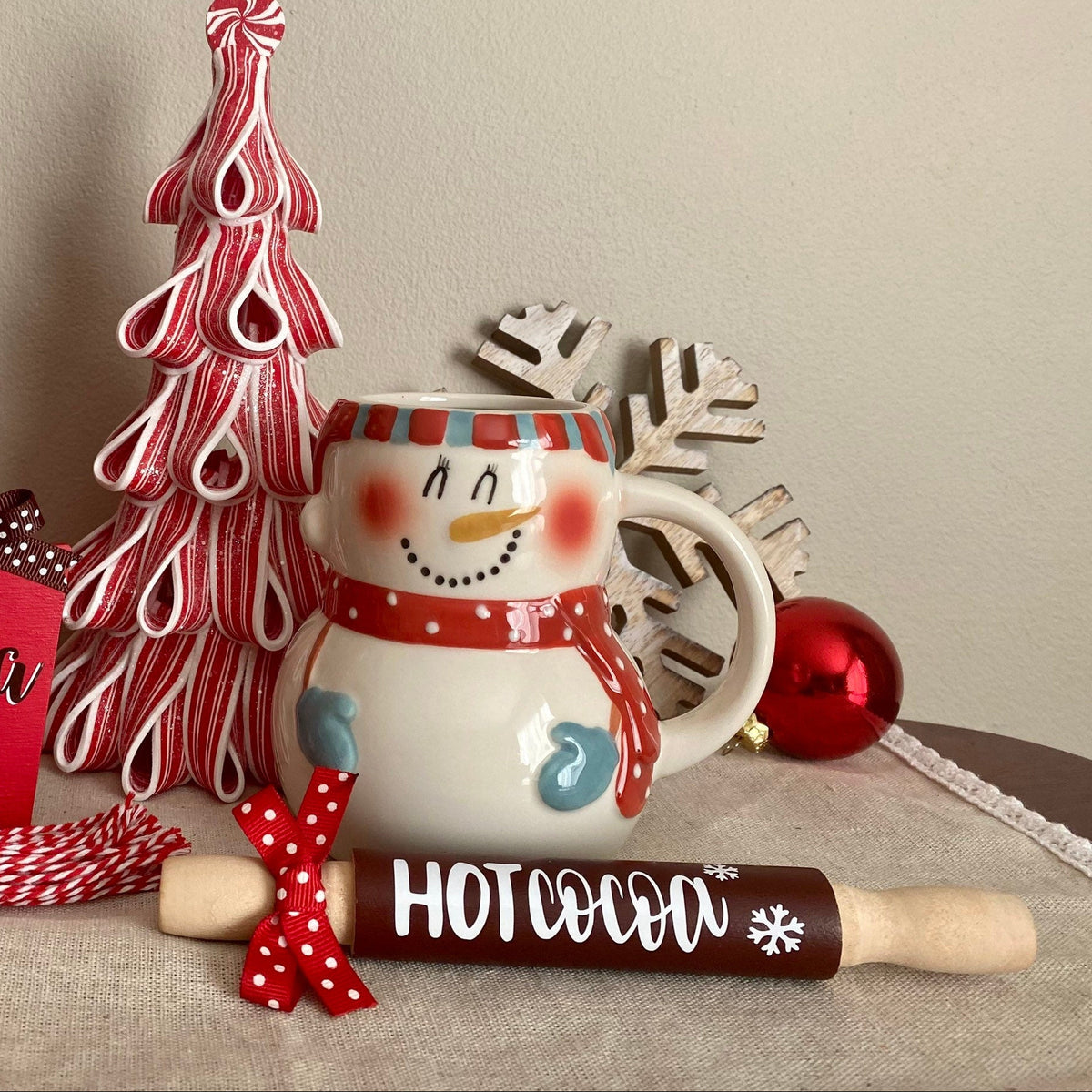 A holiday-themed hot cocoa gift set arranged on a tabletop against a plain beige wall. The set includes a tall stack of red cups with white snowflakes, a red and white striped decorative tree, a snowman mug, a red heart-shaped tag reading 'HOT COCOA', a red-and-white striped candy cane, a red-and-white striped rolling pin, and a red ornament.