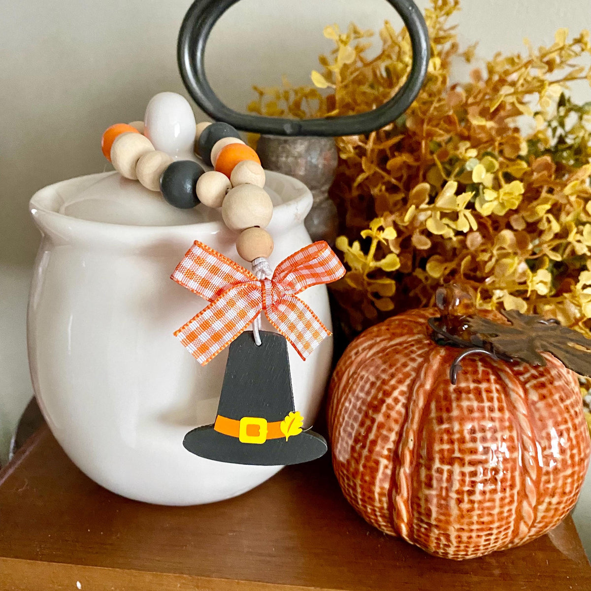 An autumn-themed wooden bead garland with orange, white, and black beads, a white gingham bow, and a black Pilgrim hat ornament with a yellow belt, draped over a white ceramic pot next to a decorative orange pumpkin.
