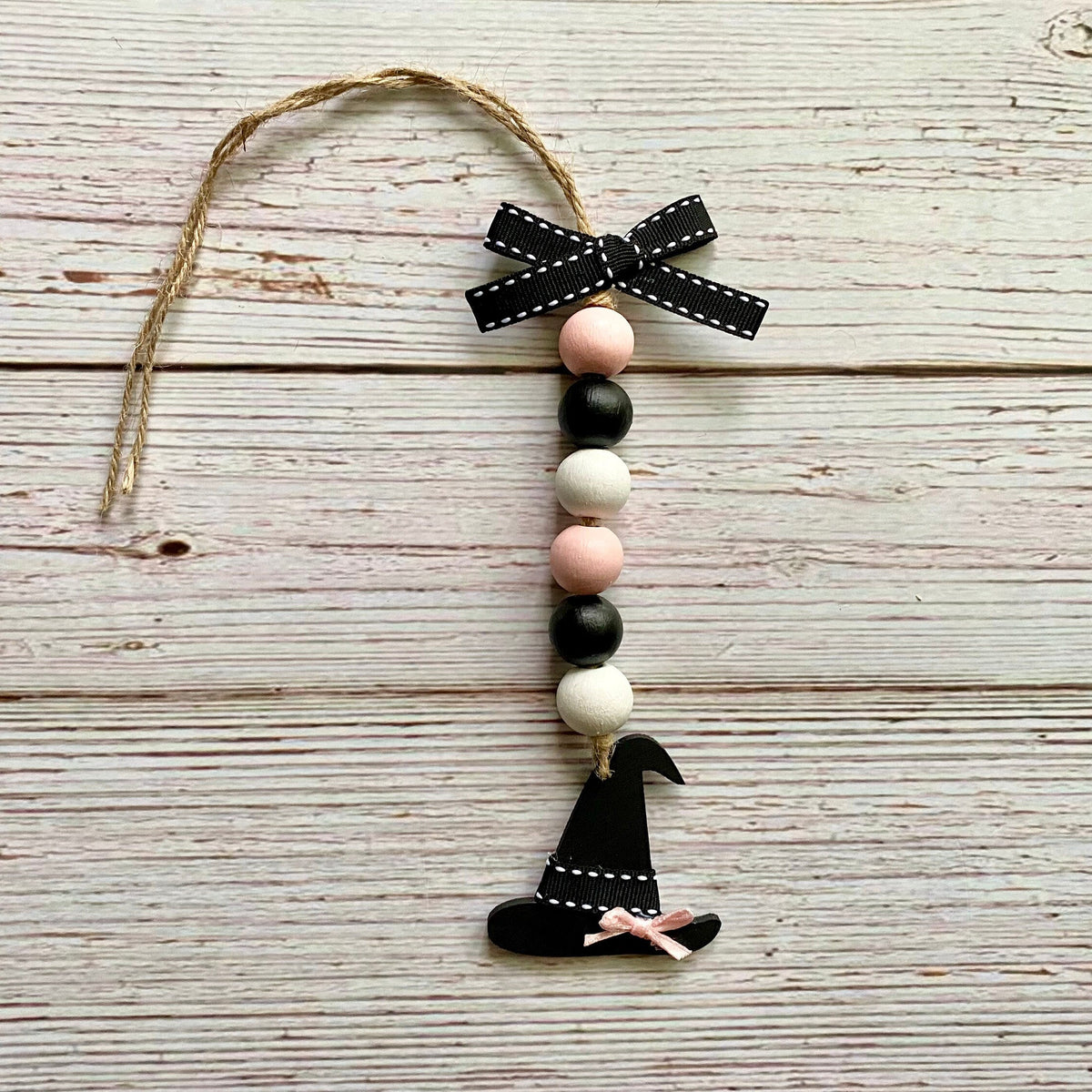 A Halloween-themed wooden bead garland ornament featuring black and white beads, two pink beads, a black witch hat, and a black ribbon bow, hanging from a jute string on a whitewashed wooden background.