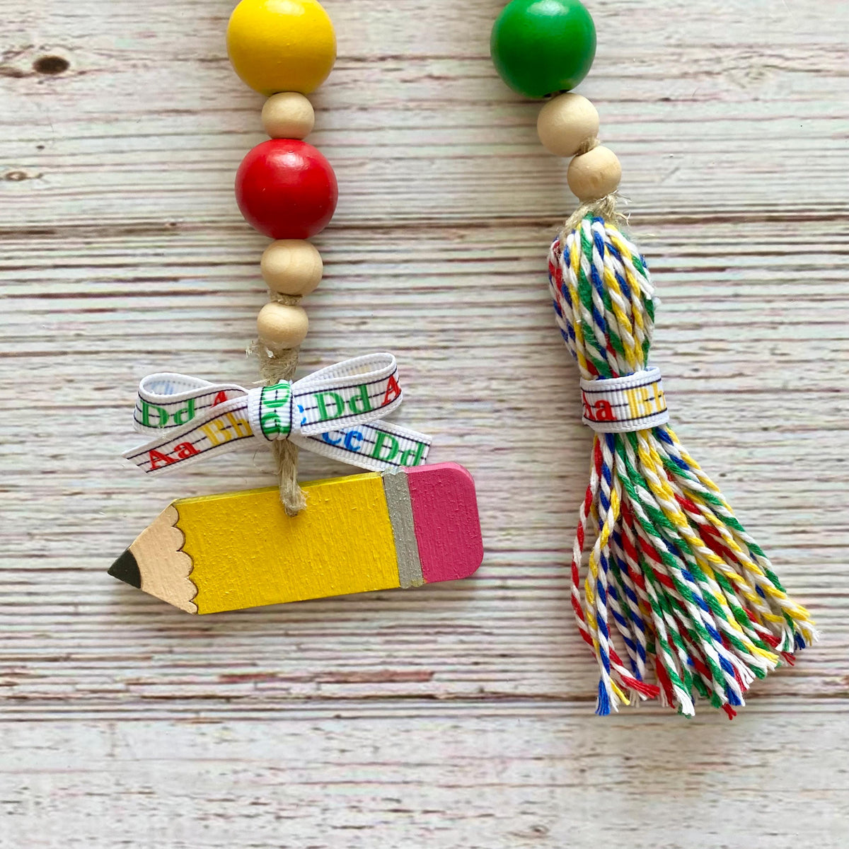 A handmade educational bead garland featuring a yellow wooden pencil charm with a pink eraser, tied with a white ribbon printed with the word 'Brick'. The left side has wooden beads in yellow, red, and natural wood, while the right side has a colorful braided tassel wrapped with a matching ribbon.